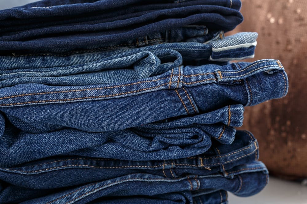 A stack of neatly folded blue jeans in various shades is placed on a flat surface, with a brown cushion in the background.